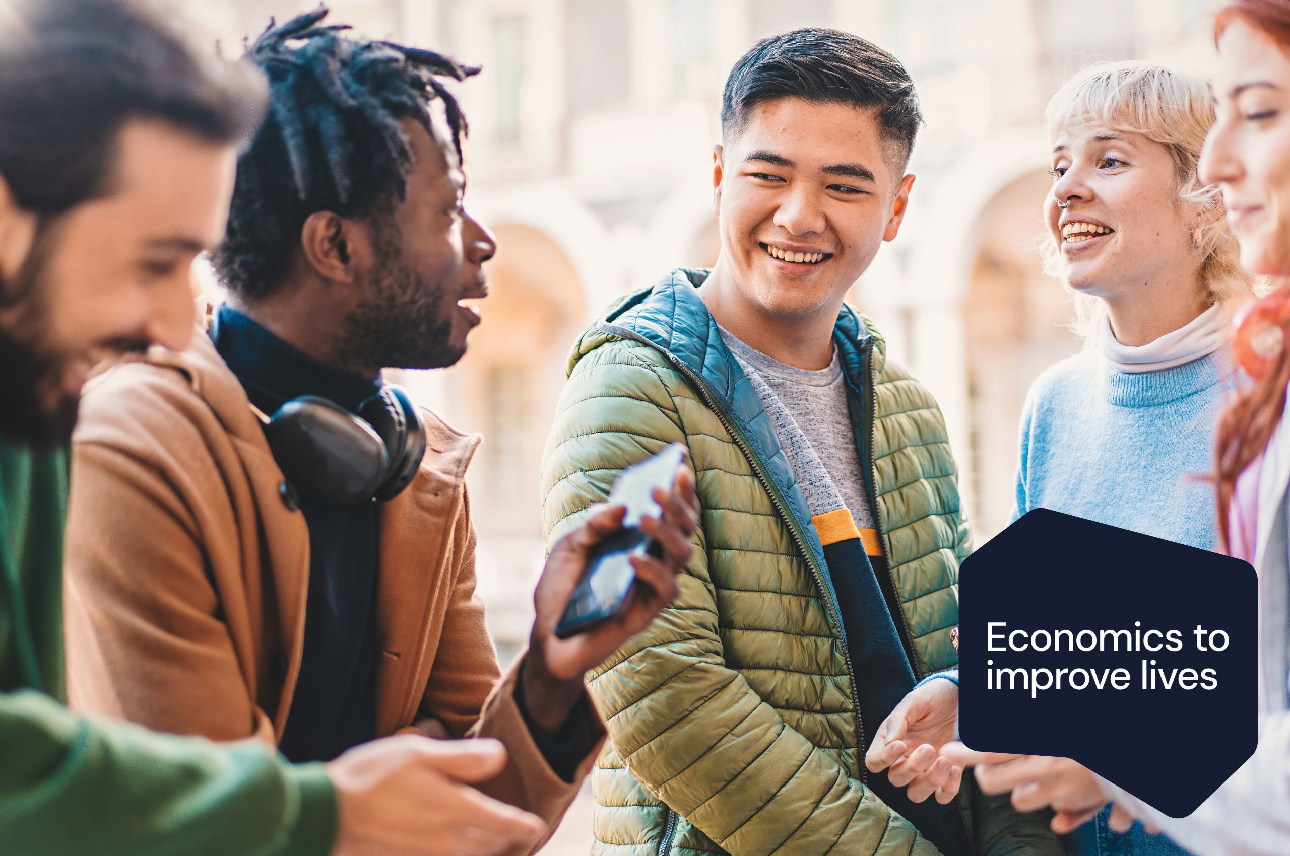 Students standing in a group chatting. There is an 'Economics to improve lives' badge in the right hand corner