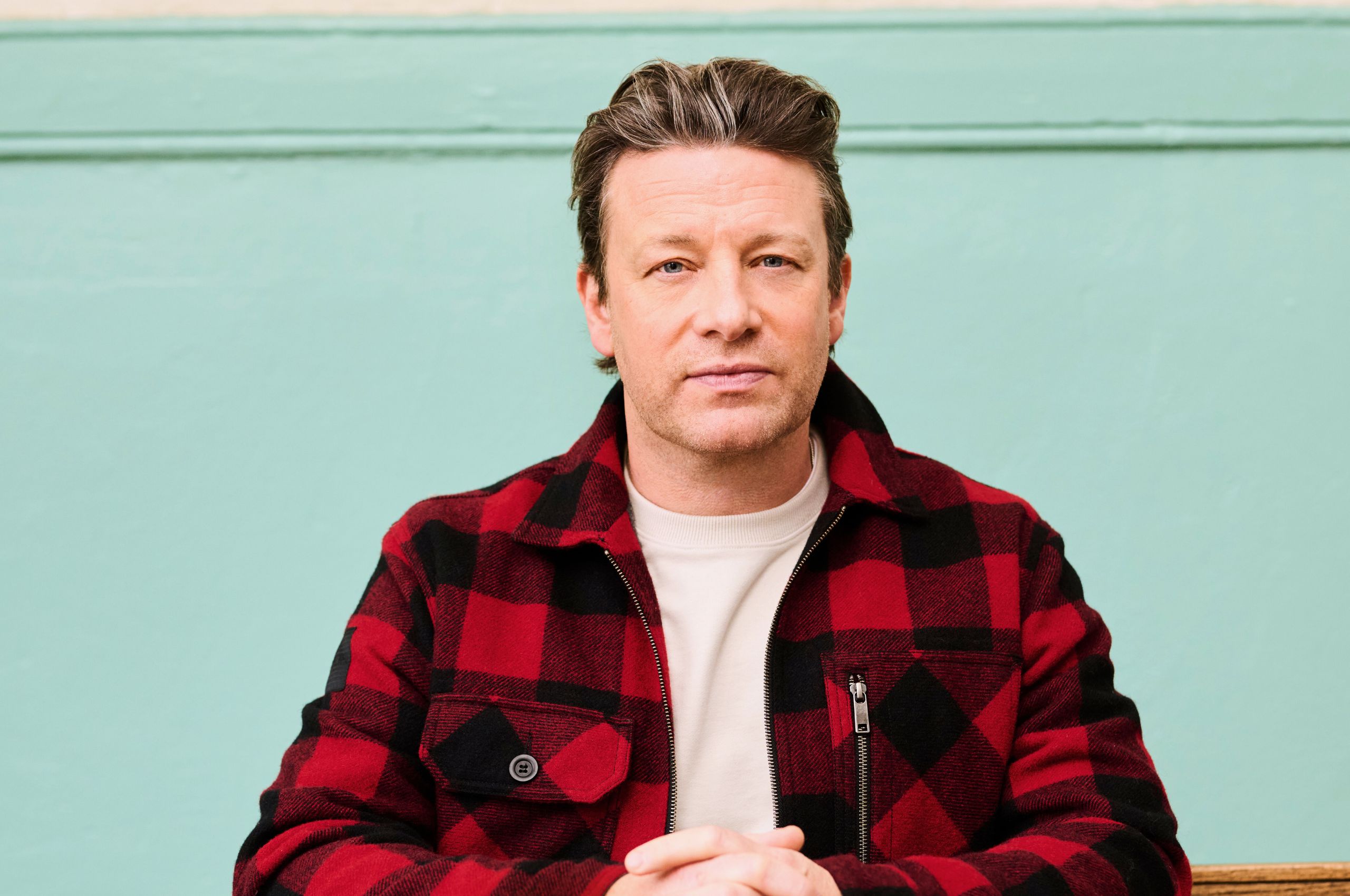Jamie Oliver, wearing a plaid shirt, sitting at a desk with his hands clasped in front of him