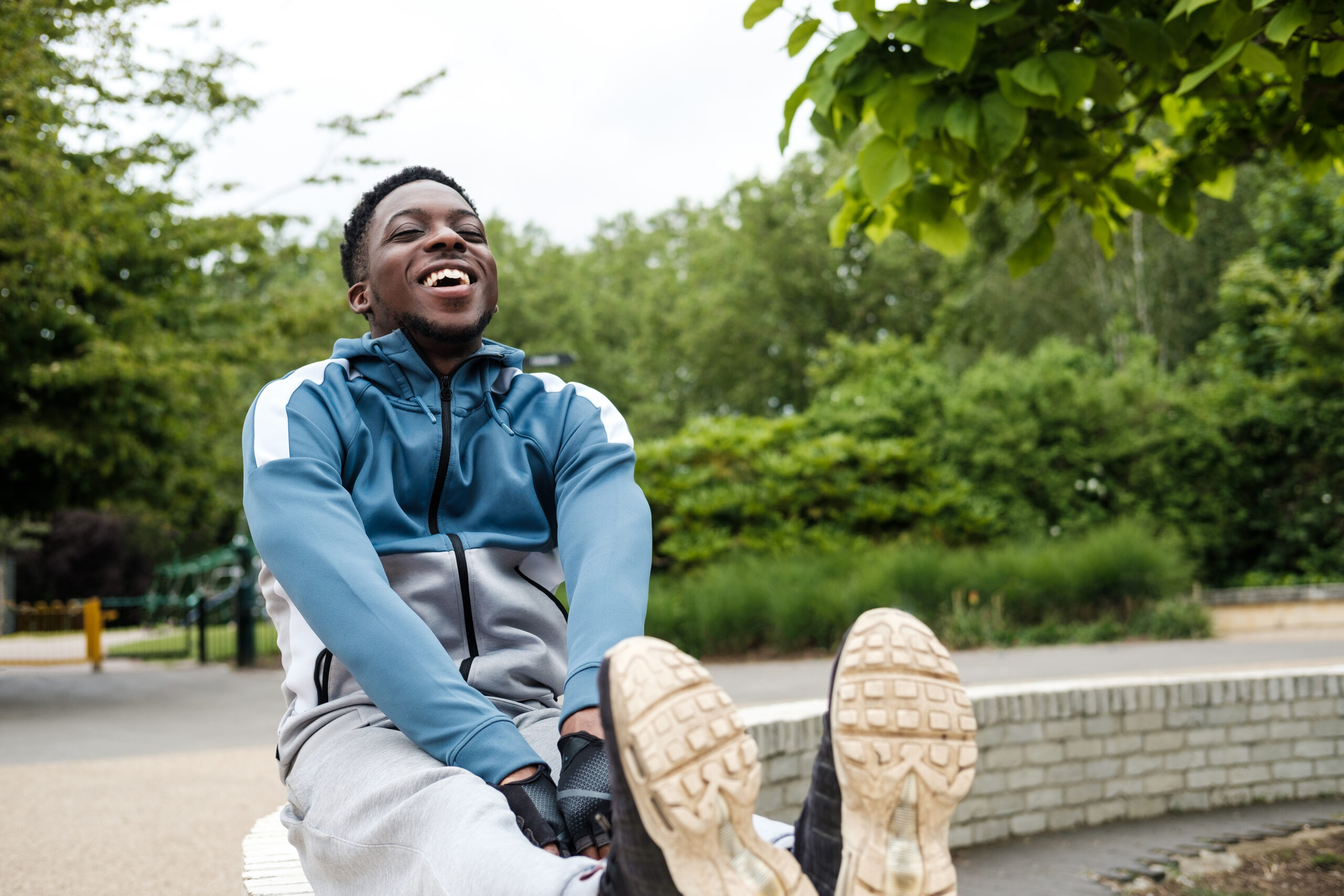 Fit young man stretching his legs in a park. He is wearing a white and blue hooded sweatshirt. He is laughing.