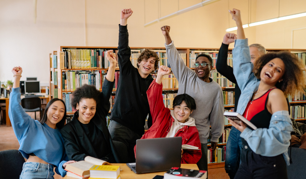 image shows young people cheering with hands in the air. They are sitting in the library around a laptop.