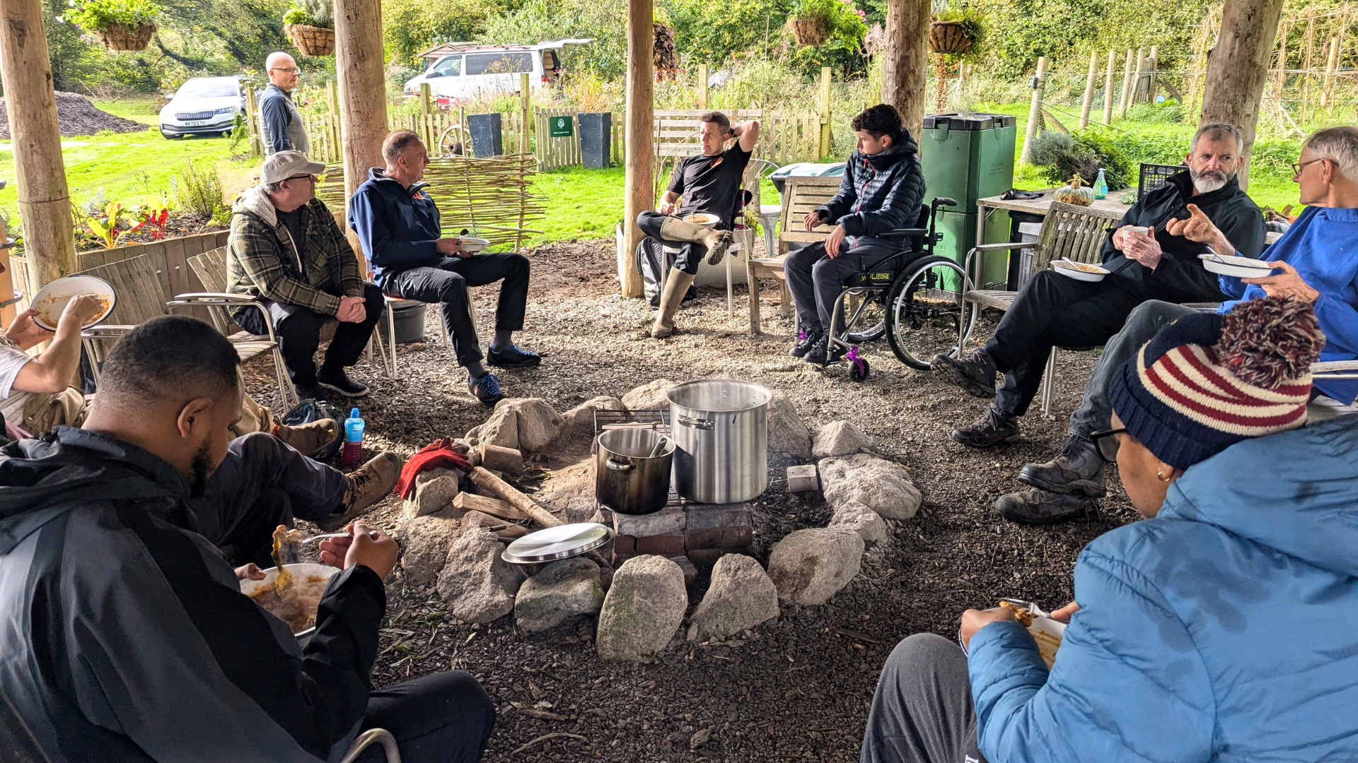 A group of veterans sit around an unlit campfire chatting and eating lunch.